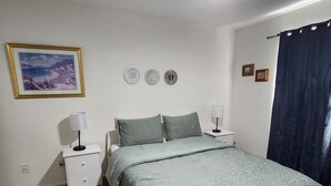 A bedroom with a bed, two bedside tables, a nightstand, a wall-mounted lamp, a framed picture, and a blue curtain. - Spacious 5BR Retreat by Trails Pet-Friendly (Parker)