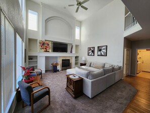 A spacious living room with a high ceiling, a fireplace, a large gray sectional sofa, and a flat-screen TV mounted above the fireplace.