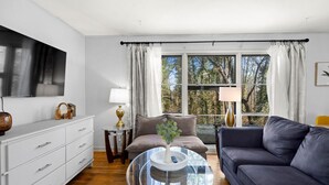 A modern living room with a flat-screen TV, a glass coffee table, a grey sofa, and a view of trees through large windows. - Cozy Home in Oak Ridge w/ 3BR (Oak Ridge)