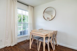 A dining area with a wooden table and chairs, a round mirror on the wall, and a window with curtains.