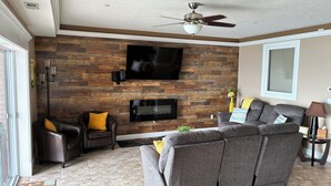 A living room with a large sectional sofa, a flat-screen TV mounted on a wooden wall, a ceiling fan, and a floor lamp. - Lakefront Condo 2br/2ba 208 indoor pool, beach/balcony. Rent 3/4th free-ask (Geneva)