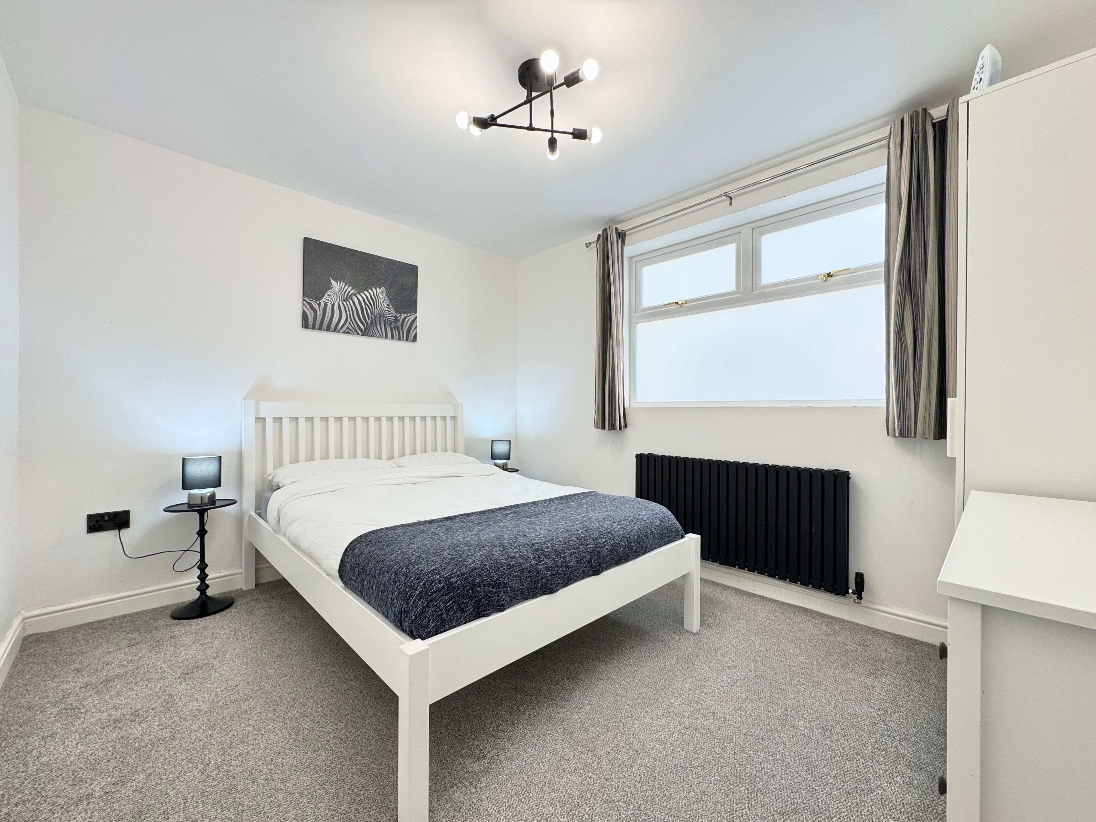 A modern bedroom with a white bed, a black radiator, a bedside table with a lamp, and a zebra artwork on the wall.
