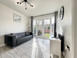 Relax in this stylish living room with a sofa, floor lamp, and flat-screen TV, offering a view of the garden through a large sliding glass door.