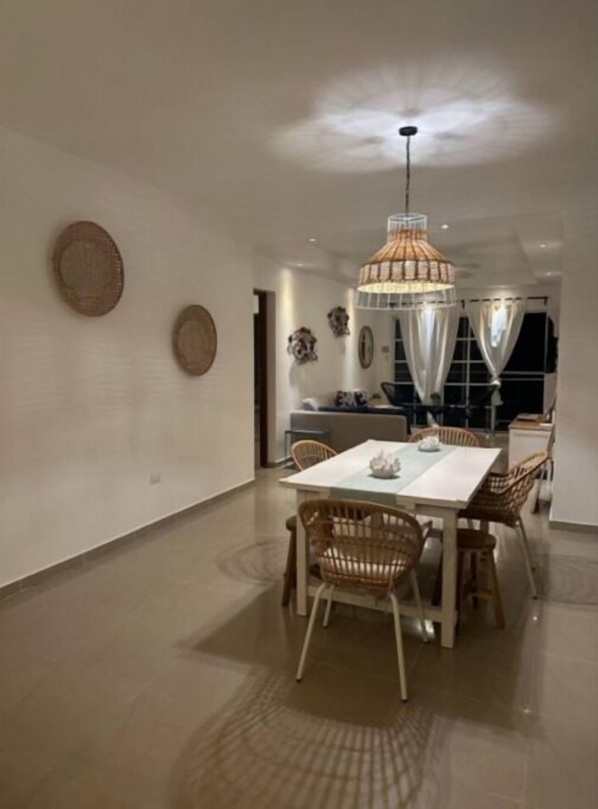 Un vaste espace dédié à la salle à manger, avec une table blanche et des chaises en osier, un luminaire suspendu et des œuvres d’art décoratives accrochées au mur.