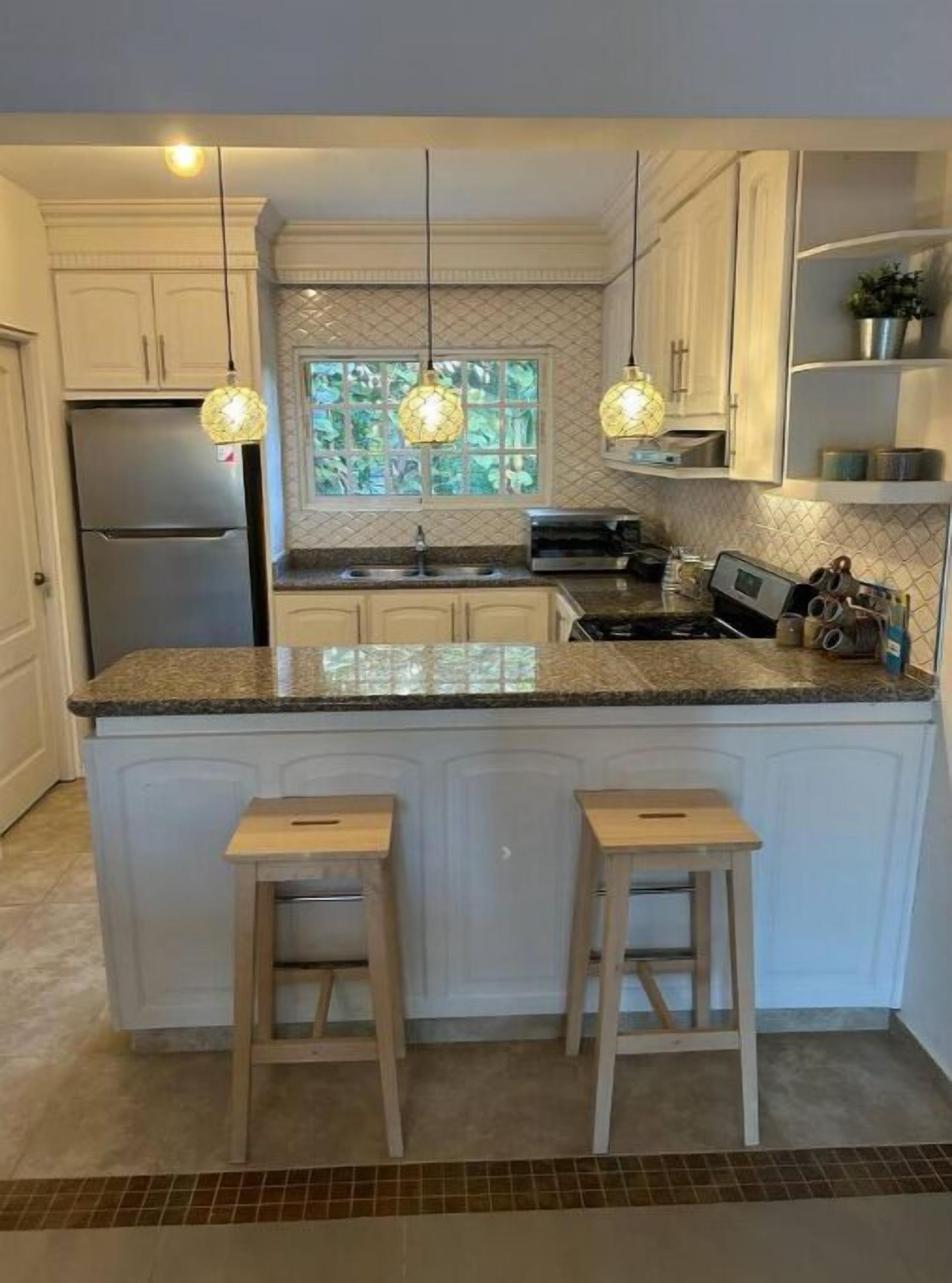 A modern kitchen with a central island, bar stools, a refrigerator, a stove, and pendant lights.