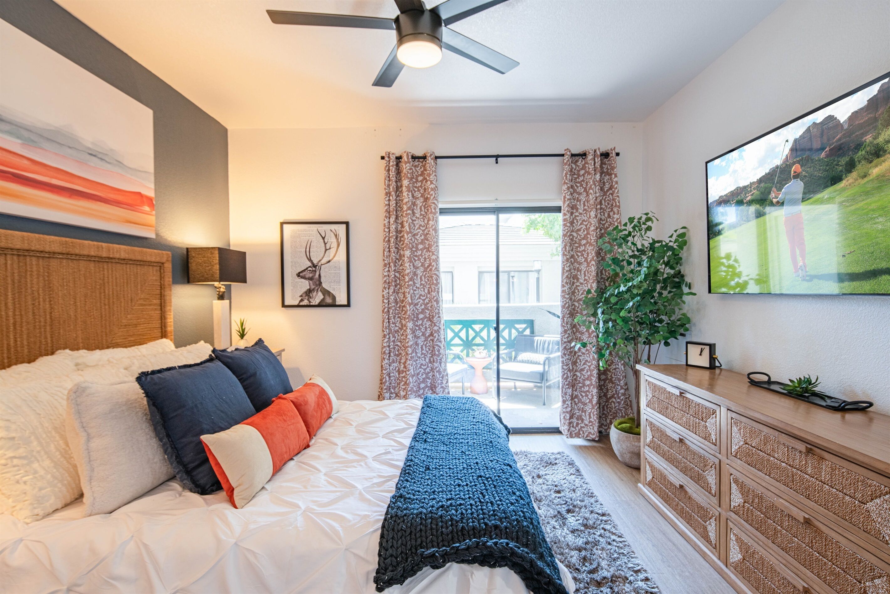 Un dormitorio con cama, ventilador de techo, ventana grande con cortinas, televisor y planta.