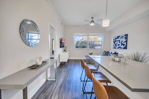 Una cocina moderna con una isla central, un comedor con sillas, un ventilador de techo y un amplio ventanal con vistas a edificios.