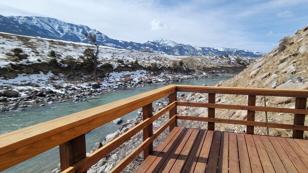 A wooden deck overlooking a river with snow-capped mountains in the background. - Yellowstone Riverside 3 Bedrooms 2.5 Bathroom Home with Panoramic Park Views (Gardiner)
