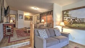 A living room with a grey sofa, patterned cushions, a red rug, a wooden staircase, and a painting of dogs on the wall. - Yellowstone Riverside 3 Bedrooms 2.5 Bathroom Home with Panoramic Park Views (Gardiner)