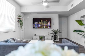 A modern living room with a cityscape TV, a white sofa, a dining table, and a ceiling fan.