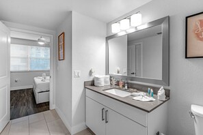 A modern bathroom with a large mirror, two sinks, and a toilet. There is a window with blinds and a door leading to another room.