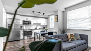 A modern kitchen with stainless steel appliances, a dining area with a gray sofa, and a large window with blinds.