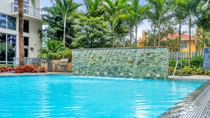 A swimming pool with a waterfall feature, surrounded by palm trees and a stone wall.