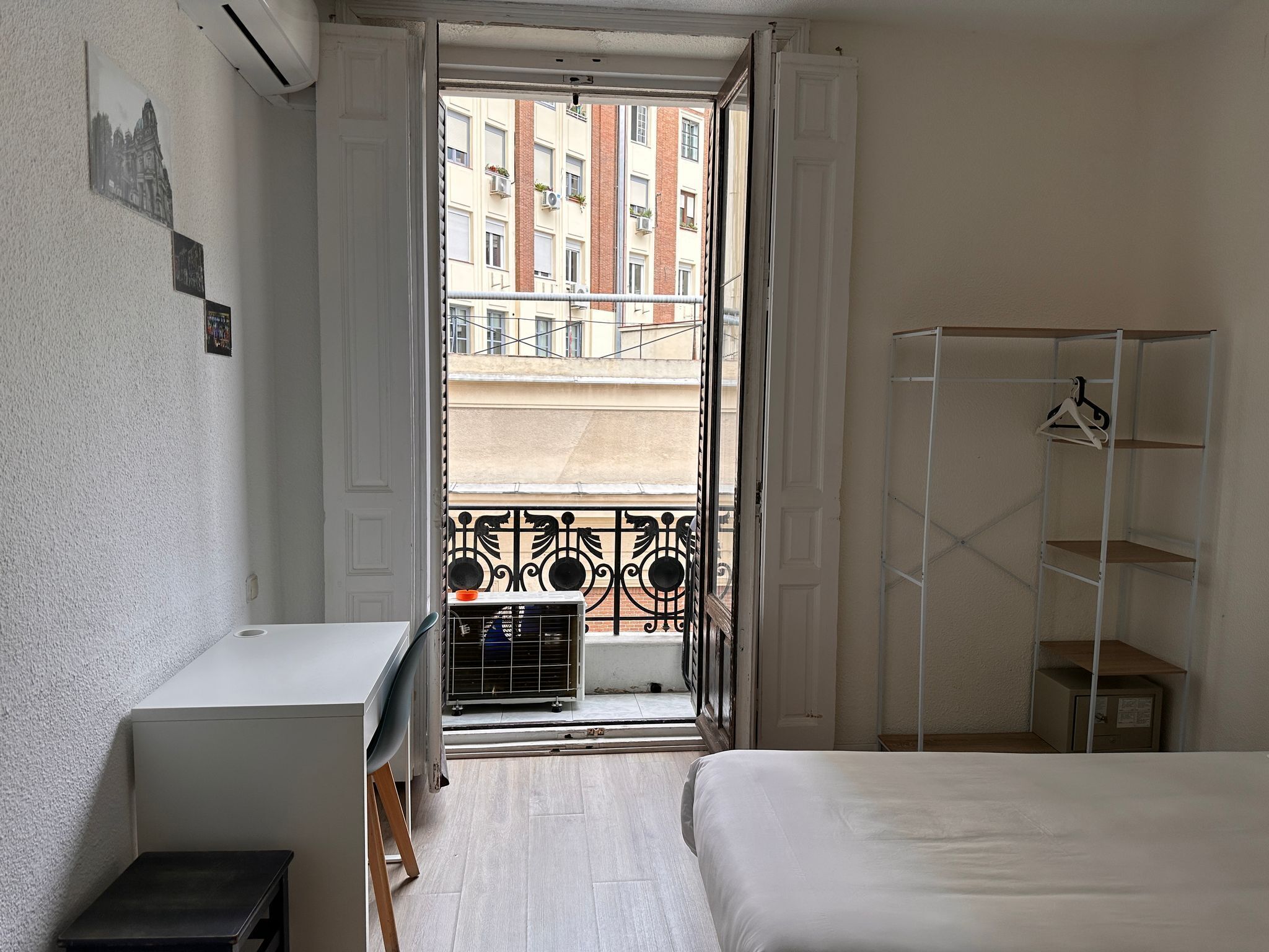 A bedroom with a bed, a desk, a wardrobe, and a balcony with a view of buildings.