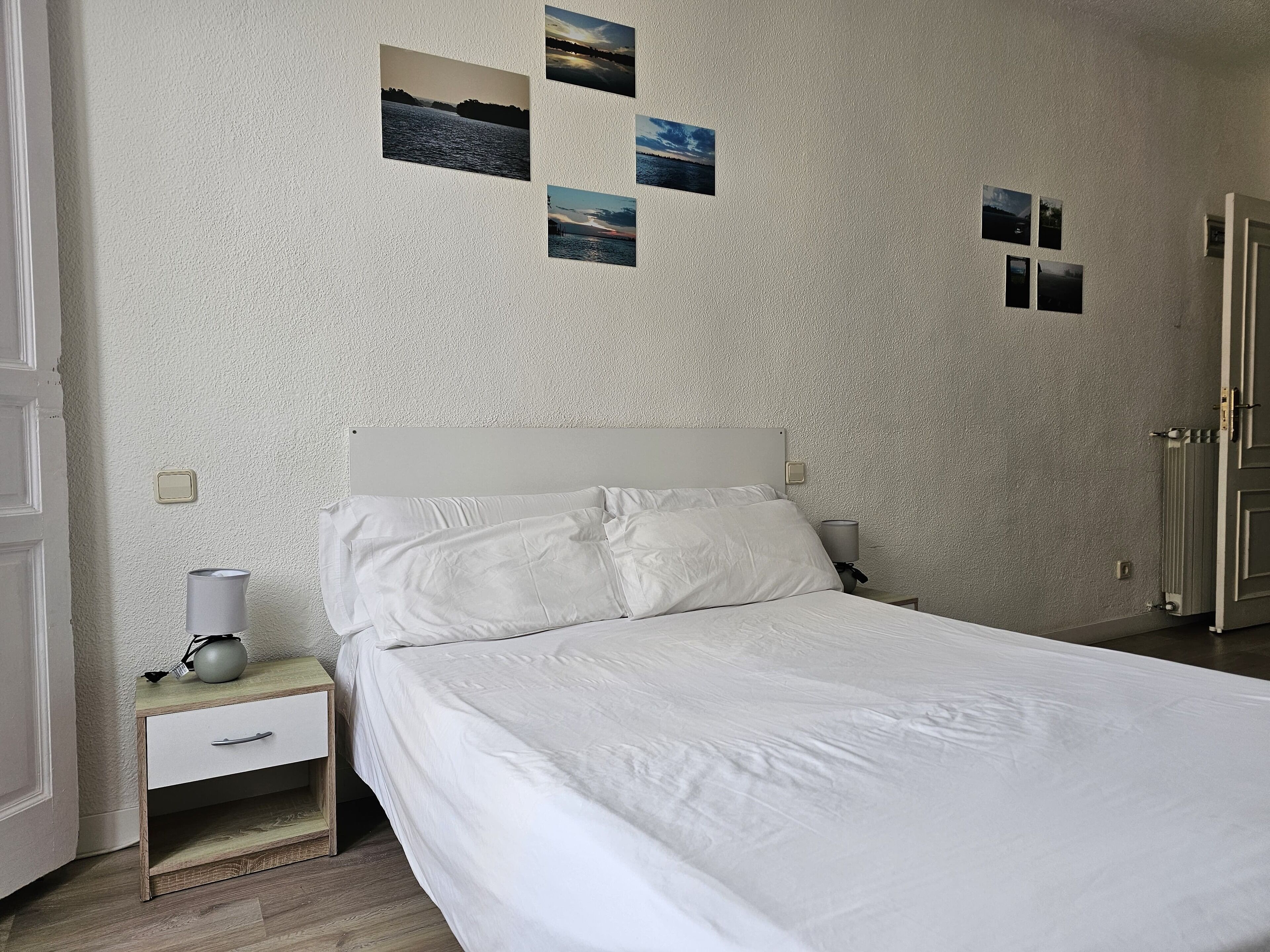 Una cama bien hecha con sábanas blancas en una habitación con pared texturizada. Hay cuatro cuadros enmarcados en la pared y una mesita de noche con una lámpara y una taza.