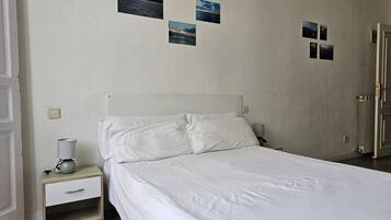 Una cama bien hecha con sábanas blancas en una habitación con pared texturizada. Hay cuatro cuadros enmarcados en la pared y una mesita de noche con una lámpara y una taza.