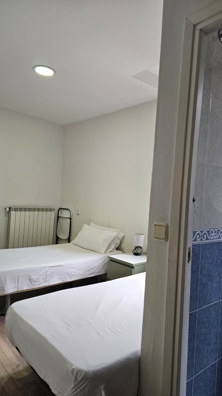 Habitación de hotel con dos camas individuales, un radiador, una mesita de noche con lámpara y una puerta con un diseño de azulejos azules.