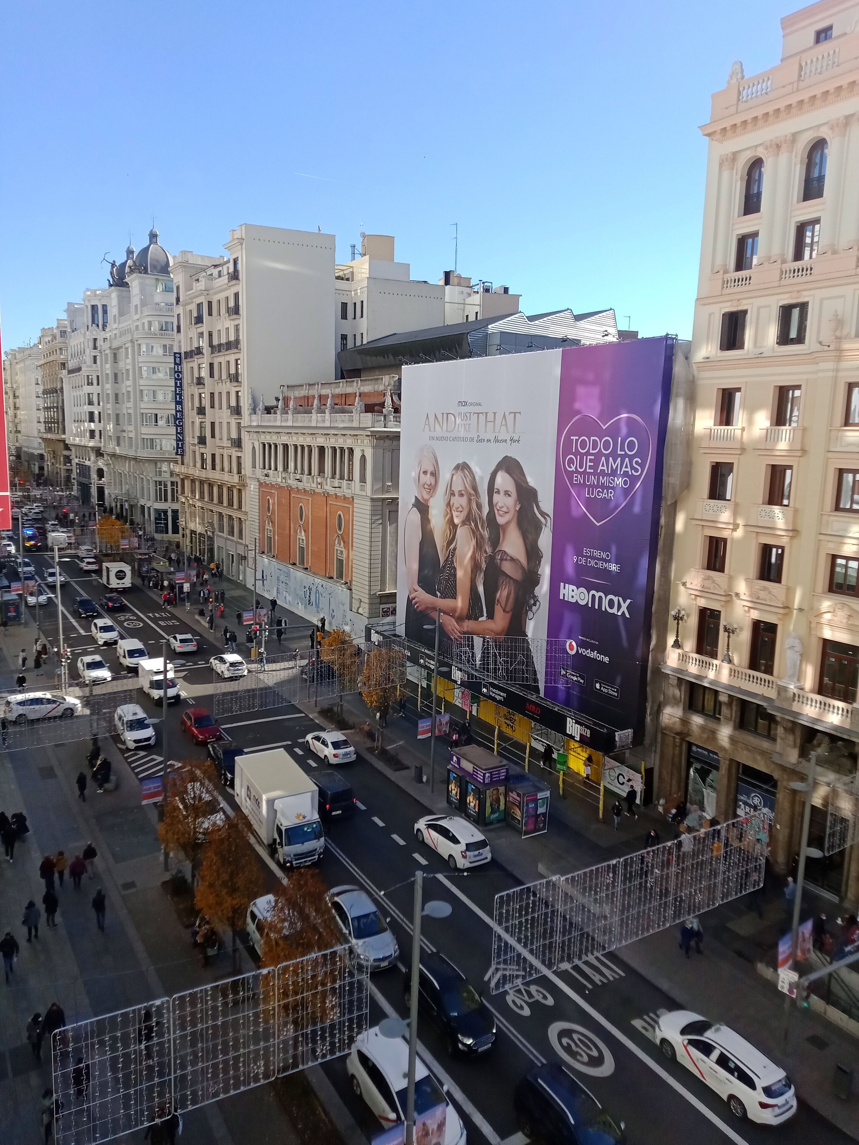 Una calle urbana concurrida con edificios históricos, un gran cartel publicitario de una película y una línea de tranvía.