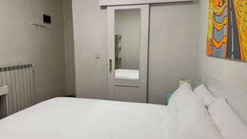 Una habitación de hotel con cama, una puerta, un radiador, un estante pequeño y un cuadro llamativo en la pared.