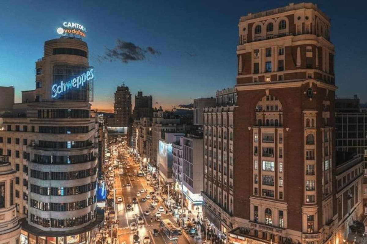 A cityscape at dusk with a prominent building labeled "Schweppes" and another with "EXITO EN MADRID" sign.