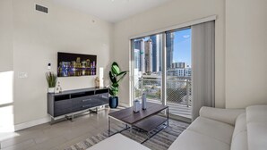 Un salon moderne doté d’un téléviseur à écran plat, d’une table basse, d’un canapé, d’une plante en pot et offrant une vue sur la ville grâce à de grandes fenêtres.