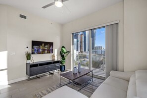 A modern living room with a flat-screen TV, a coffee table, a sofa, a potted plant, and a city view through large windows. - Skyline Views + Balcony | 2BR near Wynwood (Miami)
