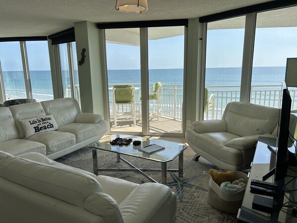 A spacious living area with a view of the ocean, furnished with white leather sofas and armchairs, a glass coffee table, and a television.
