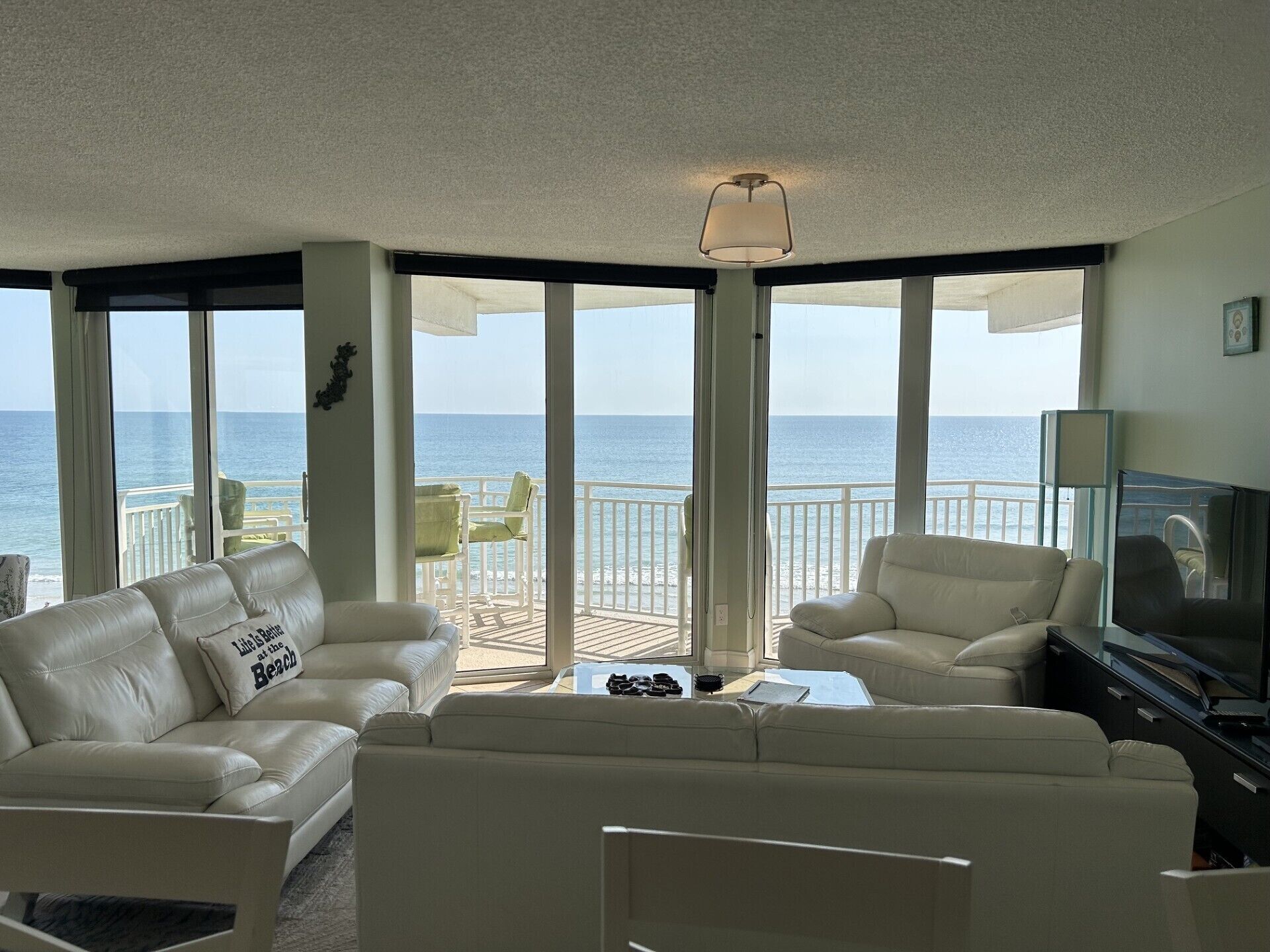 A spacious living area with a view of the ocean, featuring a white leather sofa, a television, and a balcony with outdoor seating.