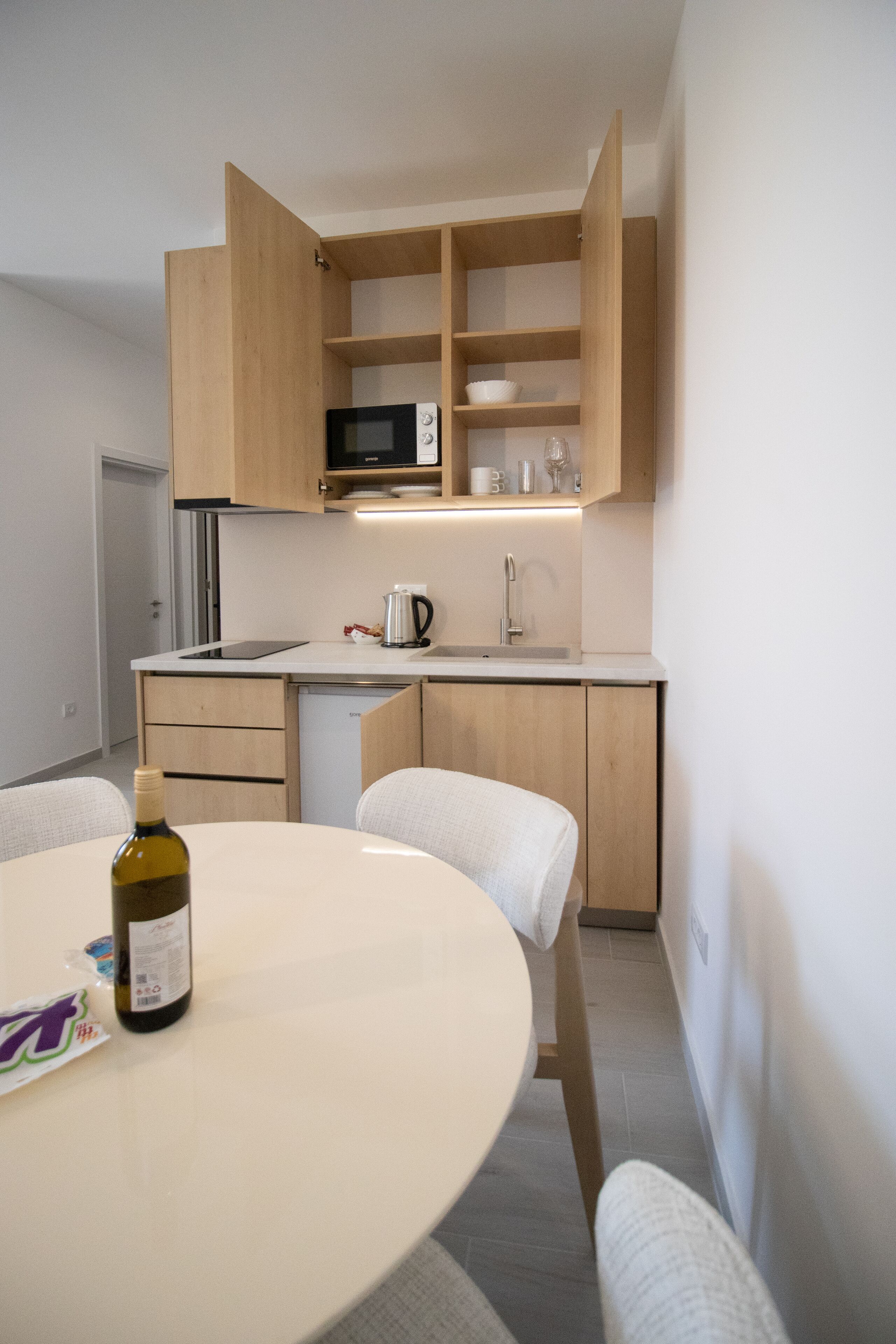 A compact kitchen with a microwave, sink, and wooden cabinets. A bottle of wine and a coffee maker are on the counter.