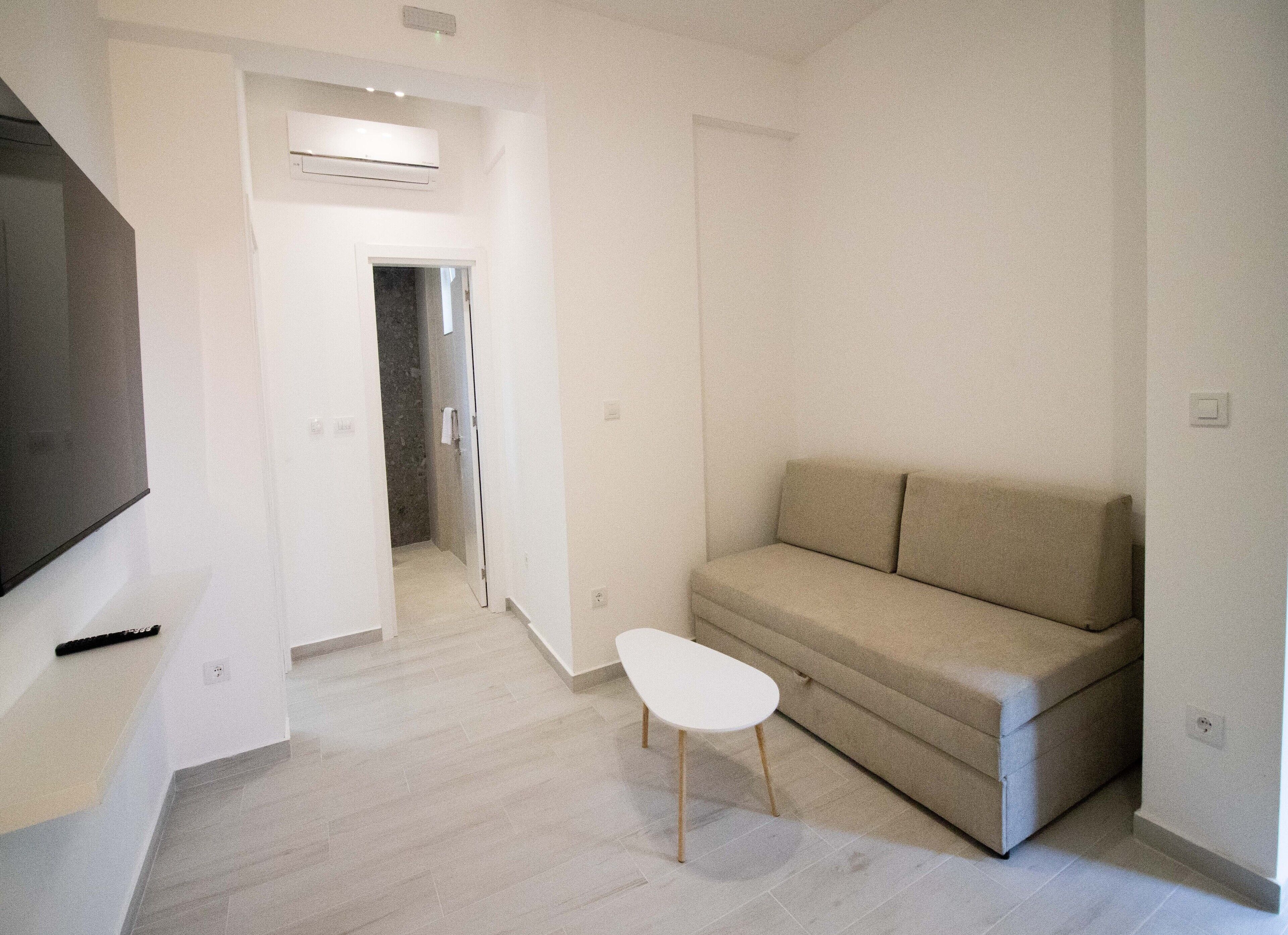 A modern living room with a beige sofa, a small white table, and a flat-screen TV mounted on the wall.