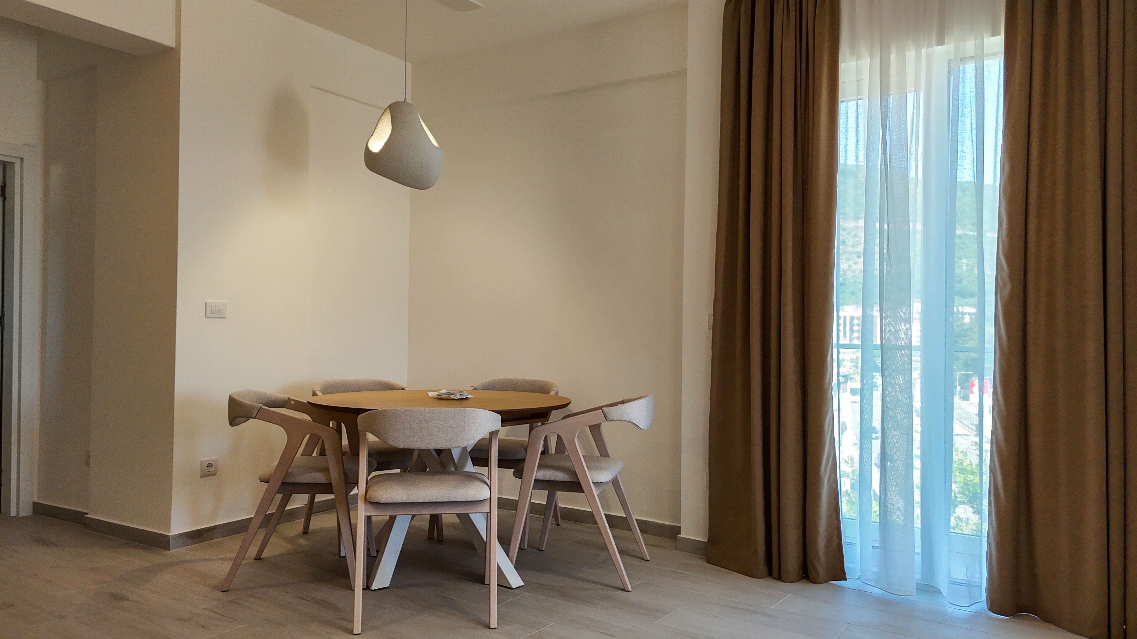 Un comedor moderno con una mesa redonda y seis sillas, una lámpara colgante y vistas a través de amplios ventanales con cortinas.