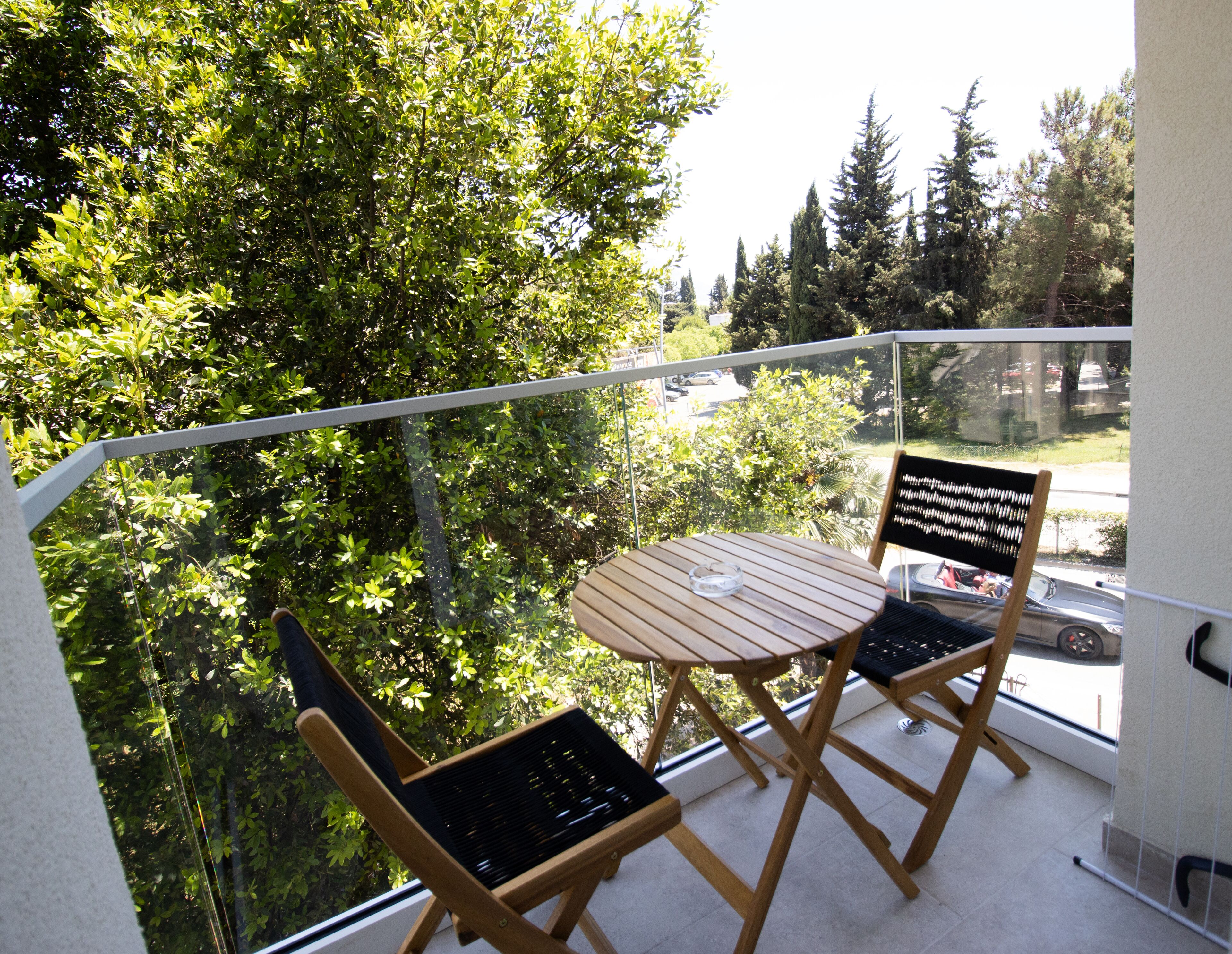 A balcony with a wooden table and two chairs, overlooking a parking area and trees.