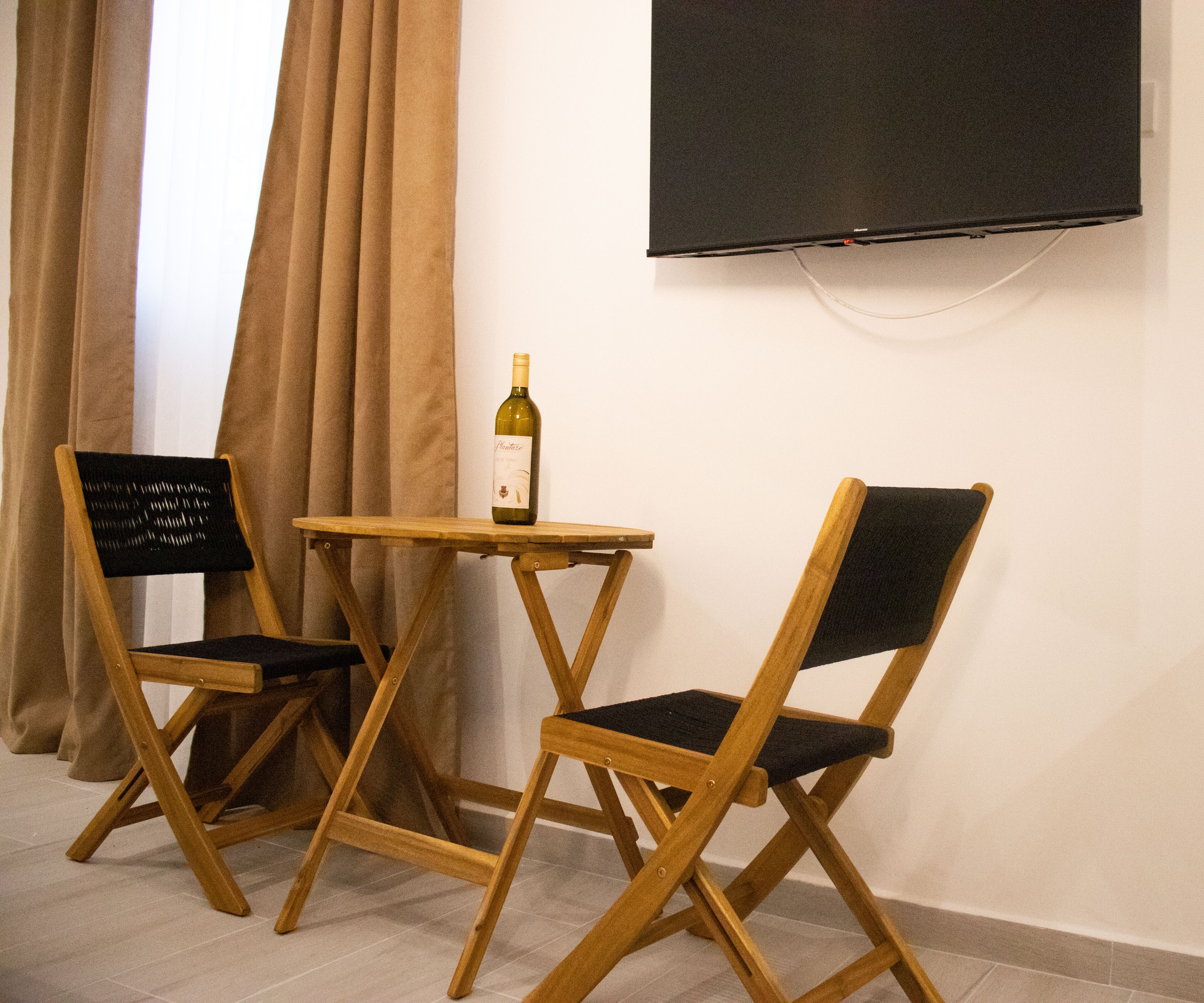 Une pièce comprenant deux chaises en bois, une petite table, une bouteille de vin et un téléviseur fixé au mur.