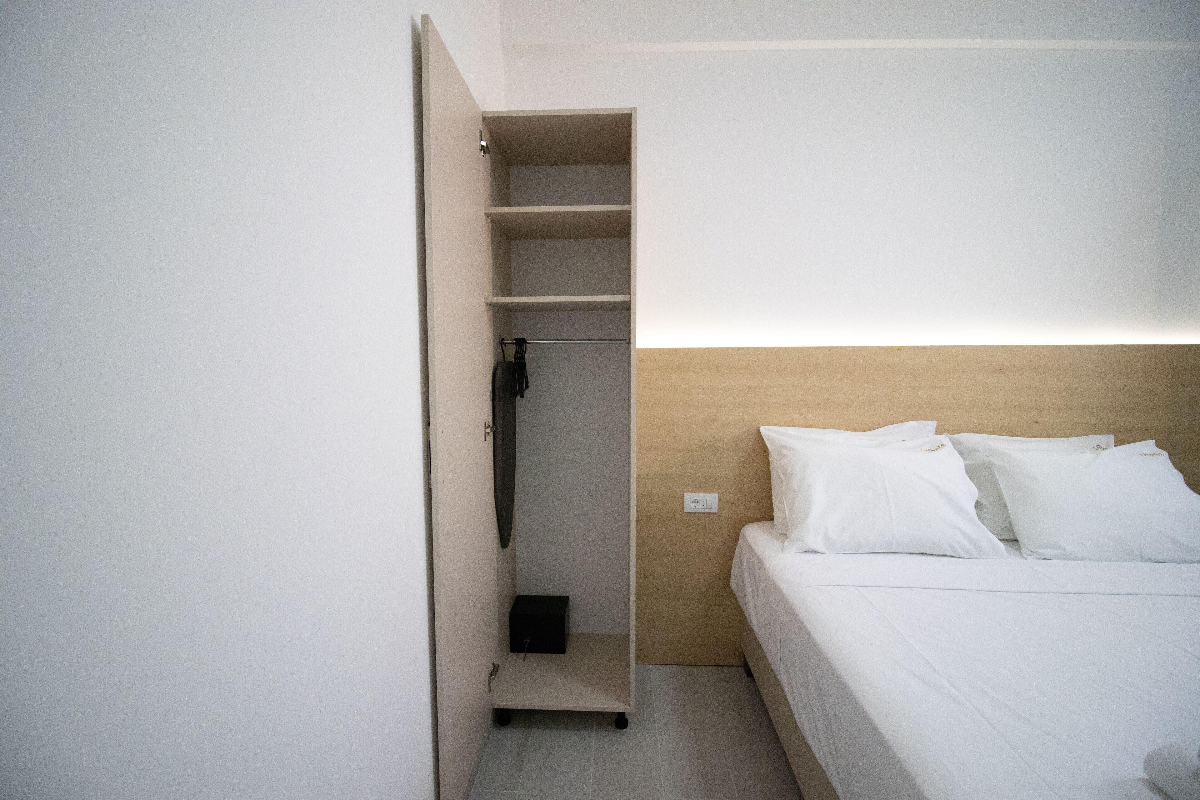 Un dormitorio con una cama, cabecera de madera, armario con repisas y una pequeña caja negra en el suelo.
