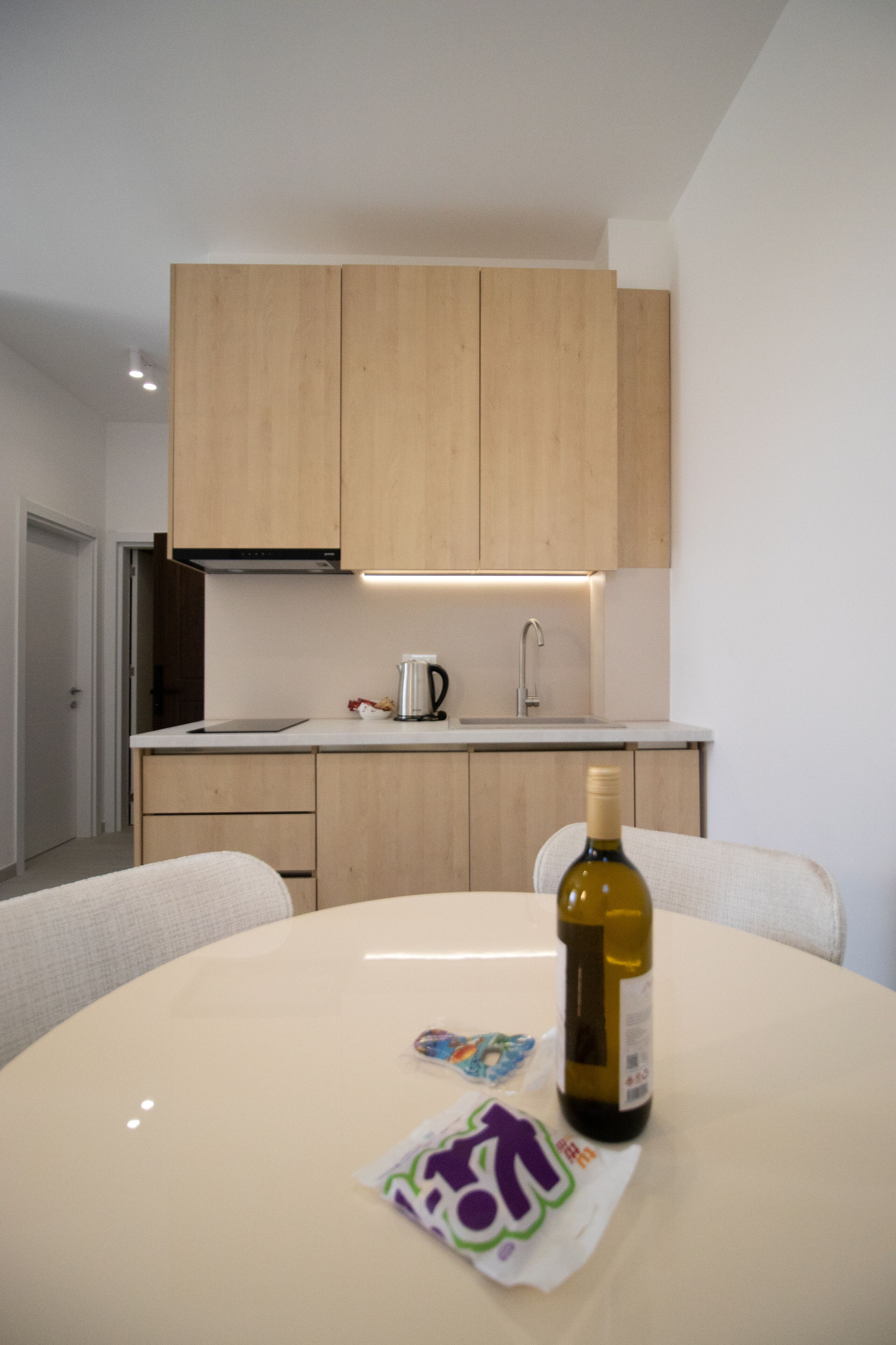 A modern kitchen with wooden cabinets, a white countertop, and a dining table with a bottle of olive oil and a packet of snacks.