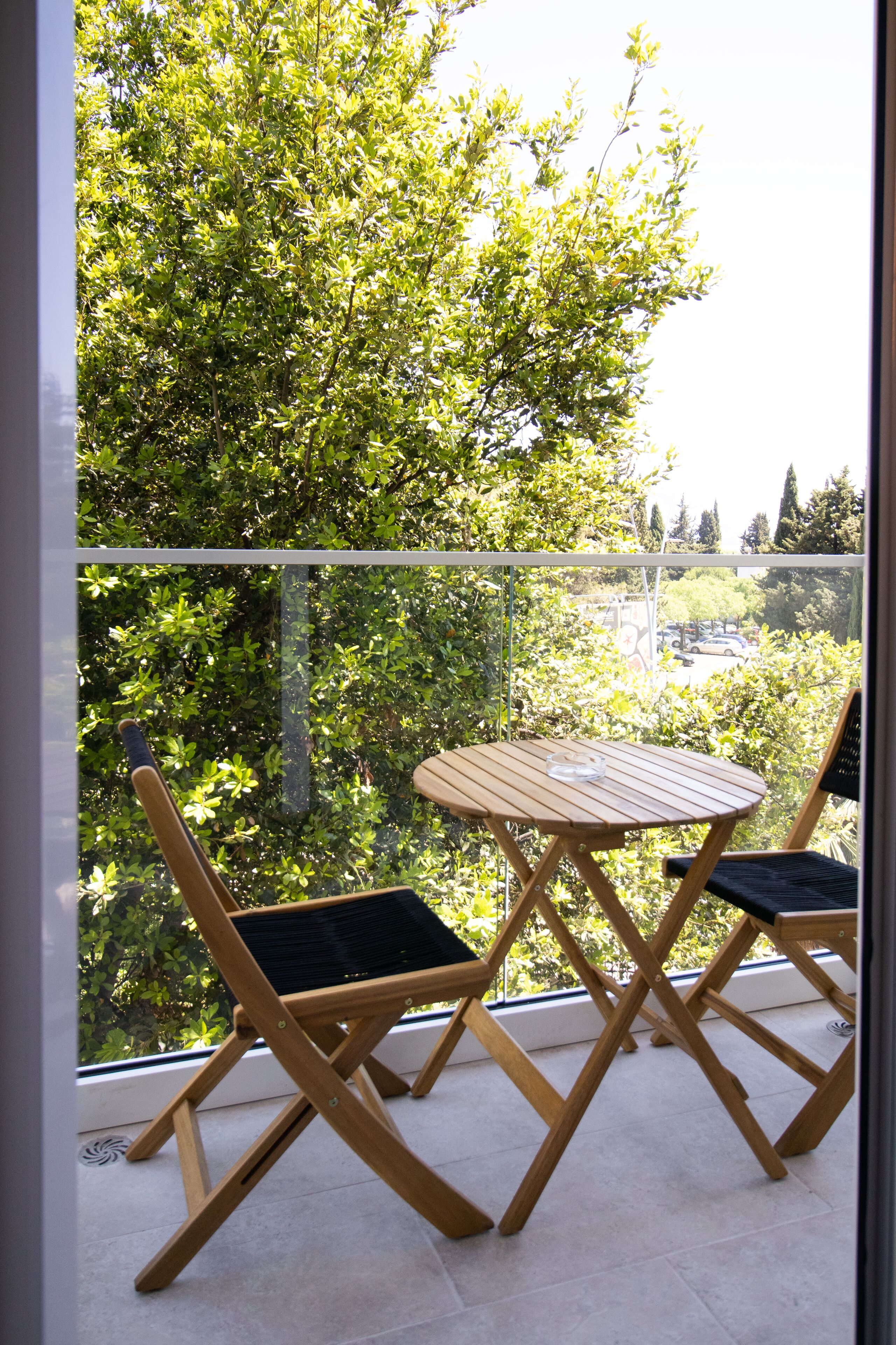 Un balcon avec une table en bois et deux chaises, donnant sur un espace verdoyant luxuriant, avec des arbres et un parking.