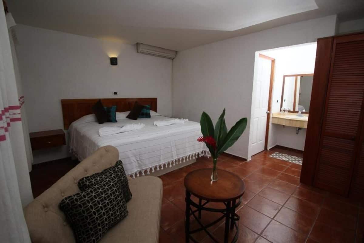 A bedroom with a bed, a bedside table, a chair, a small round table with a vase, and a bathroom visible through an open door.