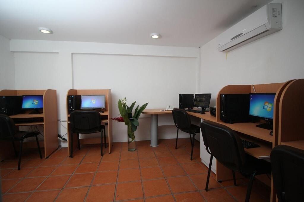 A computer lab with four workstations, each with a monitor, keyboard, and chair. There is a white table with a vase of flowers in the center.