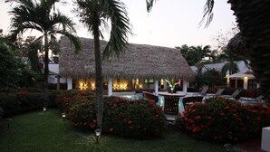 A thatched-roof building with a pool and lounge chairs, surrounded by lush greenery and red flowers. - HOTEL VILLA BLANCA SUITES (San Juan Bautista Tuxtepec)