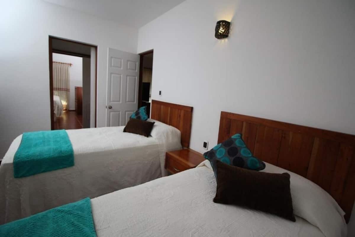 A room with two beds, each with a wooden headboard and white bedding. There is a nightstand between the beds, and a wall-mounted light fixture above the headboard on the right.