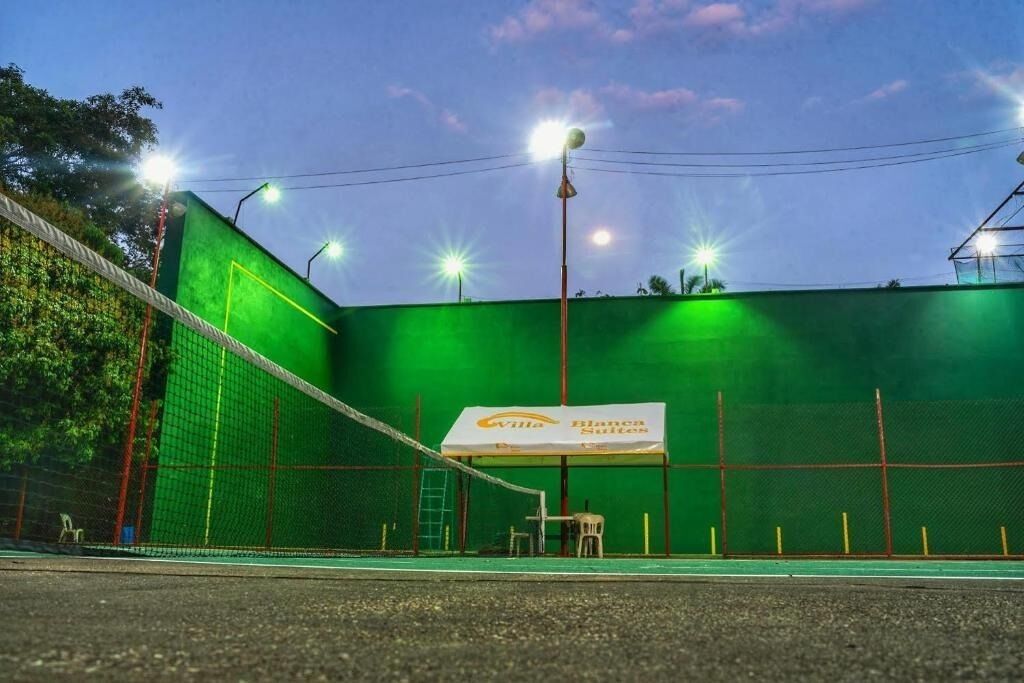 Una cancha de tenis con una red, rodeada por una cerca verde, y un letrero que dice 'Villa Blanque Suites'.