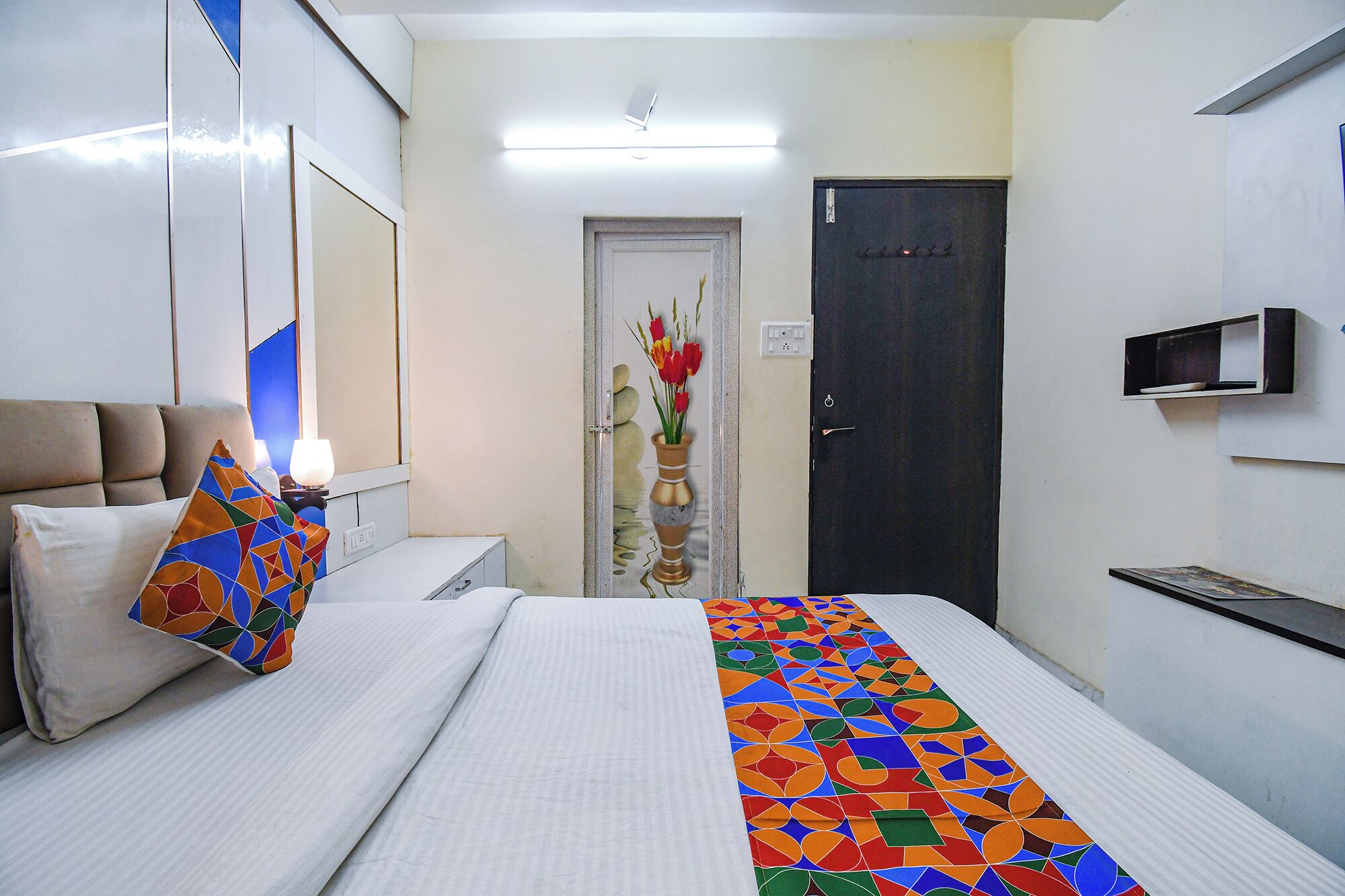 A hotel room with a bed, a nightstand, a lamp, a wall-mounted TV, and a door with a decorative vase.