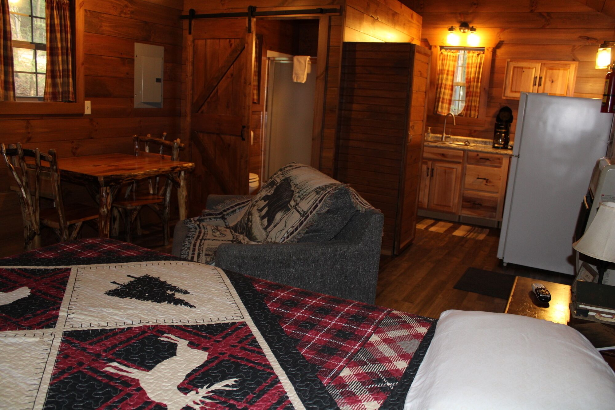 A cozy cabin interior with a dining area, a sofa with a blanket, a bed, and a kitchen with wooden cabinets.