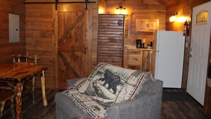 Interior acogedor de una cabaña con una mesa de comedor de madera, un sofá con una manta, una zona de cocina con armarios y un refrigerador, y una puerta de madera con mecanismo corredizo.