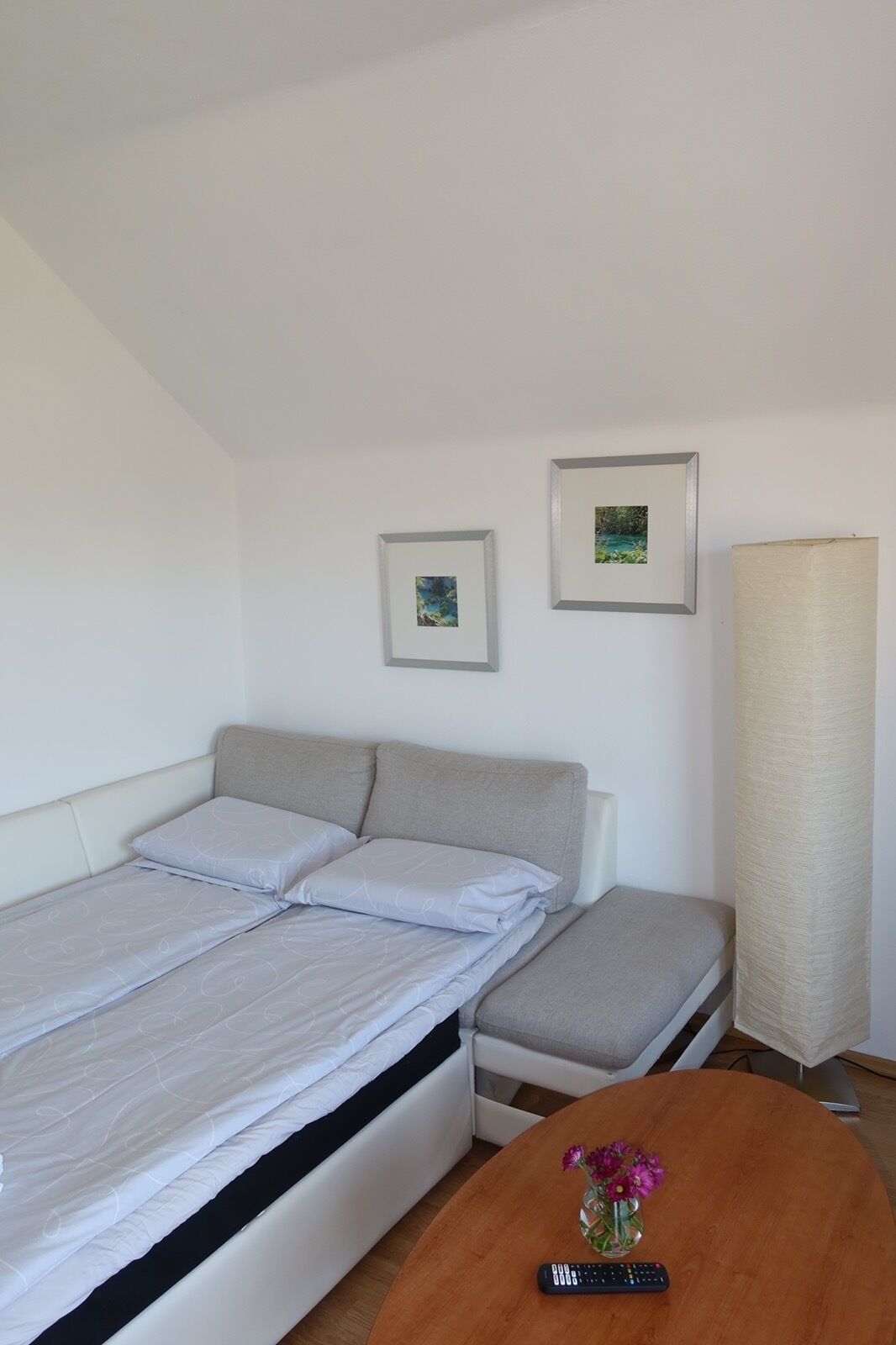 A bedroom with a bed, a bedside table with a vase of flowers, a lamp, and two framed pictures on the wall.