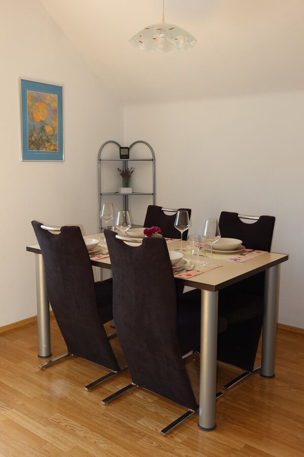 Un comedor con una mesa dispuesta para cuatro personas, cuatro sillas, una estantería metálica con un reloj y un cuadro en la pared.