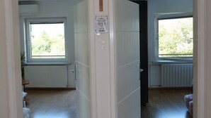 Et værelse med trægulv, hvide vægge og et bjælkeloft. To vinduer giver naturligt lys, og en dør fører til et andet værelse.