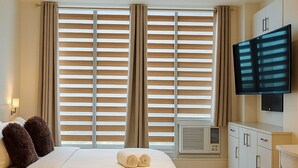 A bedroom with a large bed, a flat-screen TV mounted on the wall, and a window with blinds. - “Modern Cozy Unit with Balcony & Fast Wi-Fi – Near SM Bicutan” (Parañaque)