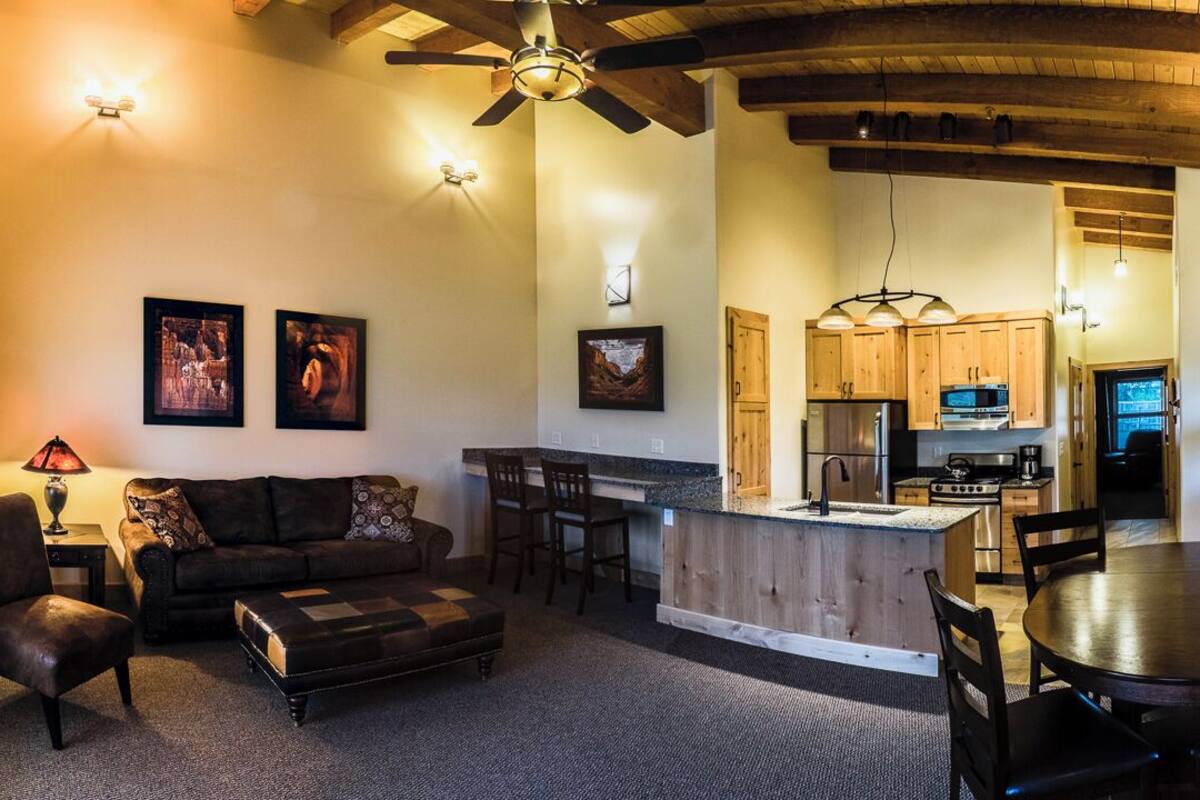 A cozy living room with a sofa, ottoman, and a dining area with a table and chairs. There is a kitchen with wooden cabinets and a central island.