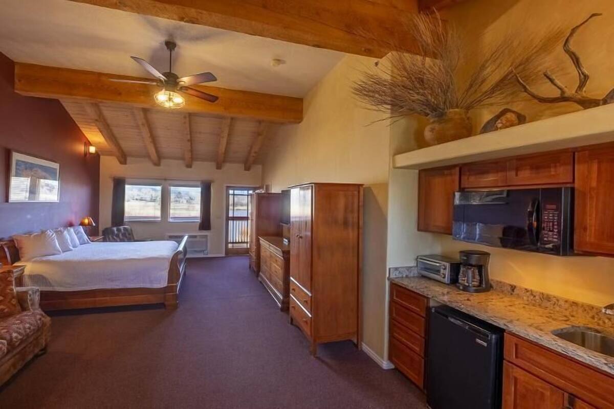 A bedroom with a bed, bedside tables, and a ceiling fan. There is a kitchen with a refrigerator, microwave, and cabinets.